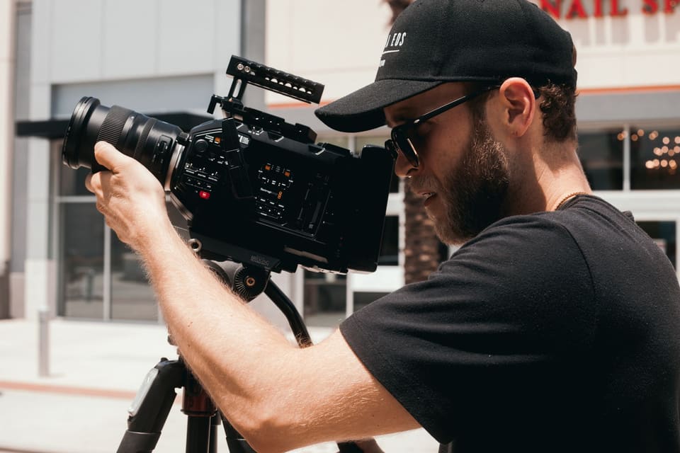 An adult male filmmaker with a beard and sunglasses operates a professional camera outdoors.